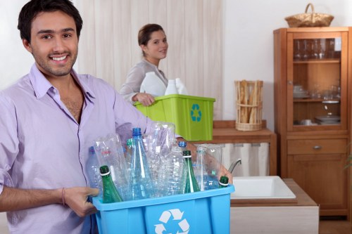 A young man with dark hair and a light purple shirt is smiling and holding a blue recycling bin filled with clear plastic bottles and aluminum cans, positioned in a domestic kitchen. Behind him, a young woman with brown hair tied back, wearing a light grey sweater, stands next to a green recycling bin with the recycling symbol, placed on a wooden countertop. The kitchen has light-colored wooden cabinets, a white sink, and a wooden display cabinet with glass doors in the background. The scene appears well-lit with natural light, suggesting a typical household environment, possibly in the Millbank area. The image is relevant to rubbish removal services provided by Garden Clearance Millbank, illustrating waste collection and recycling practices in a residential setting.