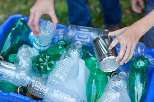 A close-up view of a person’s hands sorting plastic waste, with one hand holding a transparent plastic bottle and the other grasping a metallic tin can, amidst a variety of discarded plastic bottles and containers in shades of clear, green, and blue. The waste is piled in a container or bin set outdoors, with grass visible in the background, suggesting a garden or yard setting consistent with rubbish removal services in Millbank. The textures of the plastic bottles range from smooth and glossy to crinkled, and the tin can has a reflective metallic surface. The image highlights the type of recyclable waste that domestic or commercial rubbish removal services like those offered by Garden Clearance Millbank often collect and dispose of, emphasizing environmental responsibility through proper waste sorting.
