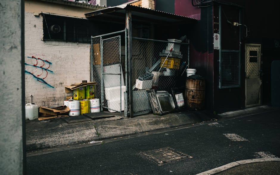 A pile of mixed rubbish and packaging materials, including flattened cardboard boxes and plastic bags, is accumulated against a rough, unpainted brick wall outdoors. The materials vary in color, prima