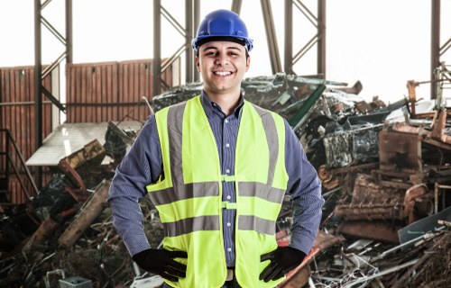 A male worker wearing a blue safety helmet, a high-visibility yellow vest, a dark grey shirt, and black gloves is standing inside a warehouse or industrial building with metal framework. Behind him, there are large piles of assorted waste materials, including rusted metal panels, broken machinery, and debris, indicating a rubbish disposal or clearance site. The environment appears well-lit with natural or artificial lighting, highlighting the details of the waste and the worker's prepared safety attire. This scene reflects a professional rubbish removal operation, similar to those provided by Garden Clearance Millbank in the Millbank area, emphasizing the handling and management of large-scale waste materials inside a structured industrial setting.